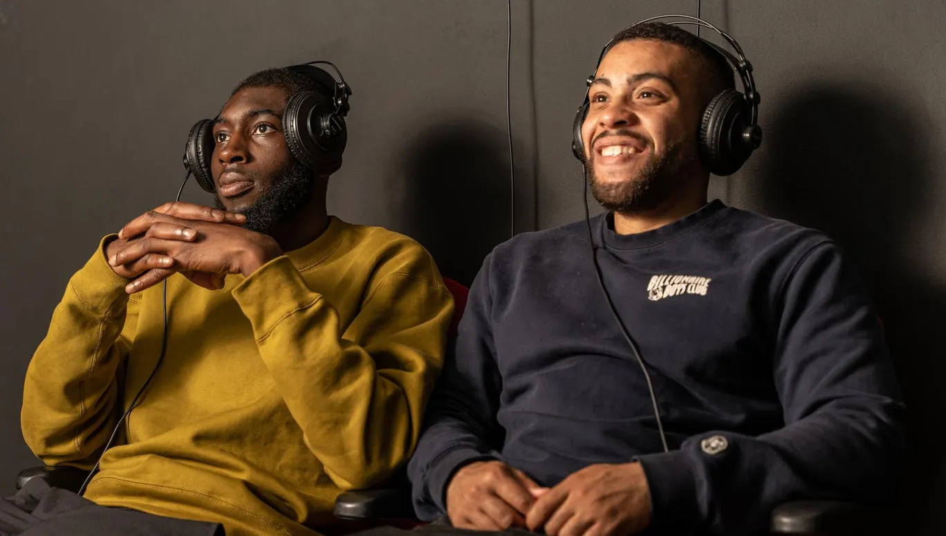 Two men wearing headphones and watching a video that is part of an exhibition at CBK Zuidoost.