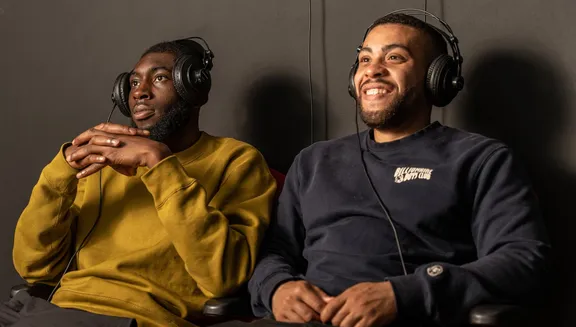 Two men wearing headphones and watching a video that is part of an exhibition at CBK Zuidoost.