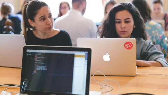 Closeup of students seated with laptops at table sat opposite each other.