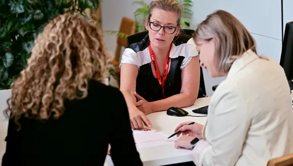Two women sitting at the desk at the INAmsterdam offices, seen from the back, one of them signing a paper. Employee seen from the front.