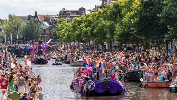 A crowd of people on the streets and in a boat with flags and signs - Pride Canal Parade