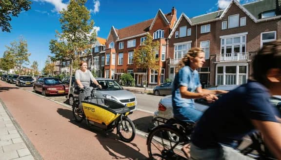 People riding bikes on Haarlem street with woman riding yellow shared Cargoroo e-cargo bike elektrische buurtbakfiets - fast commuting on Dutch street