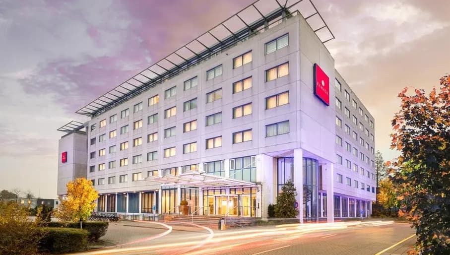 Exterior shot of Ramada Amsterdam Airport Schiphol, square building with time-elapsed trails of car lights on road outside.