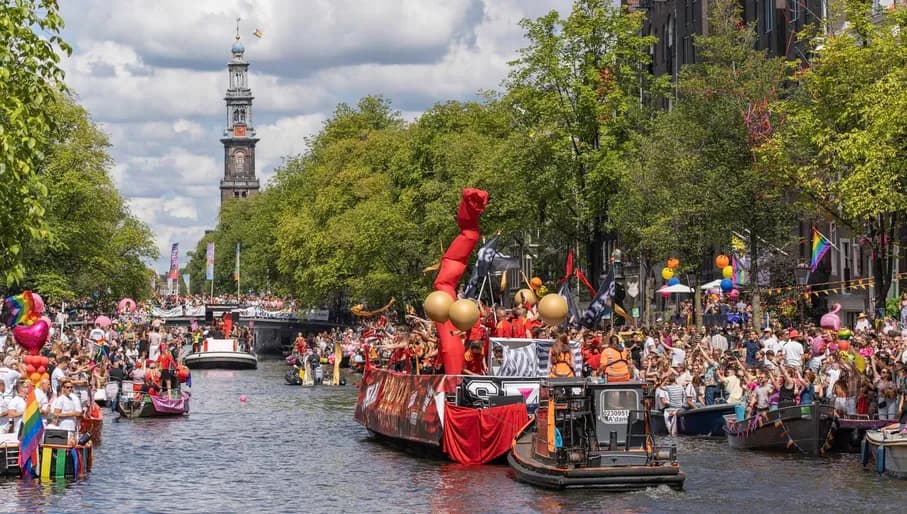A crowd of people on the streets and in a boat with flags and signs - Pride Canal Parade
