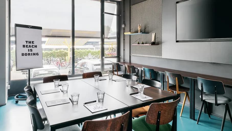 Interior of Social Hub Amsterdam West meeting room with chairs and presentation board with "The beach is boring" written on it, in front of large window showing parking spaces outside.