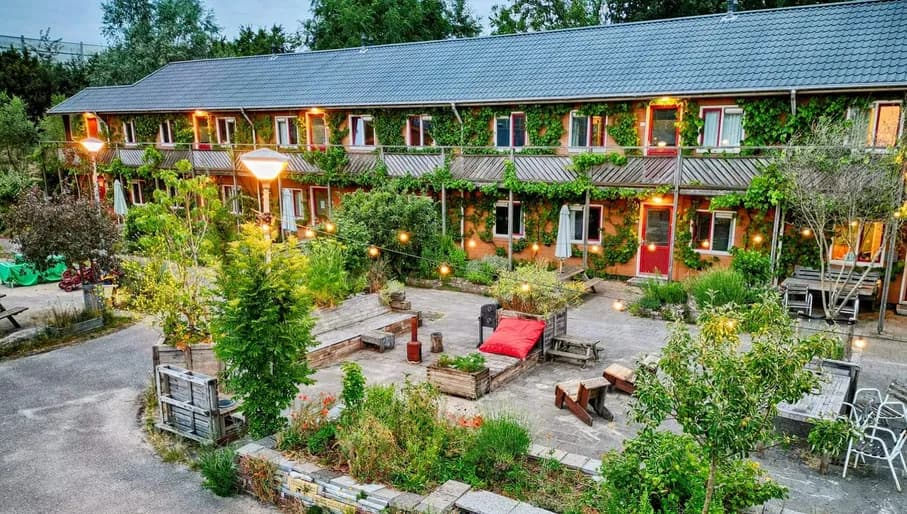 Exterior image of Amsterdam Farm Lodge at dusk, showing a two-story building and terrace with a lot of plants and lighting.